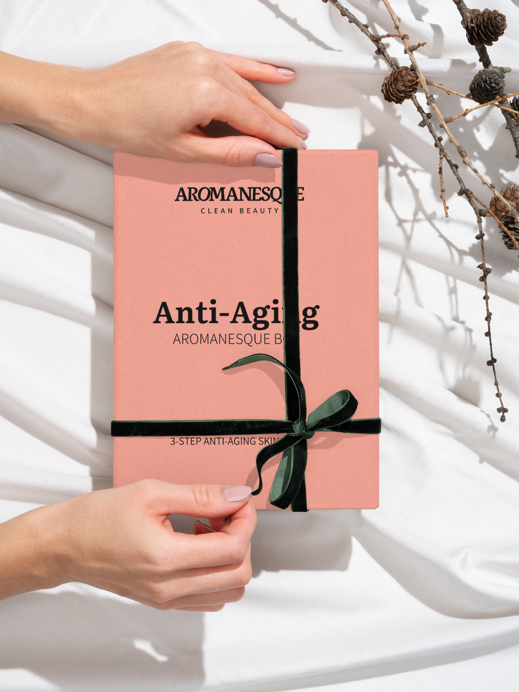 A hand holding a pink book titled 'Anti-Aging' by Aromanesque with a decorative bow, placed on a white surface with a background of dry flowers.